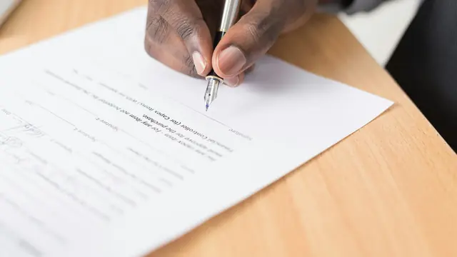 A person signing a printed document with a pen at a wooden desk